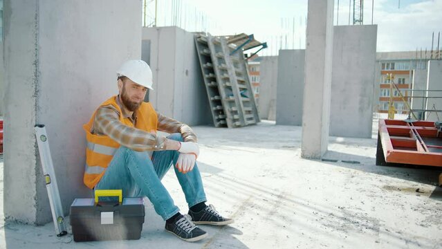 Young foreman sitting outdoors and feeling tired after overwork. Look at the camera. Caucasian worker in reflective vest and helmet at construction site. Slow motion