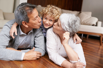 Floor, portrait or grandparents kiss a happy kid playing or hugging with love or care in family home. Smile, grandma or old man relaxing or bonding to enjoy time with child or cute boy in retirement