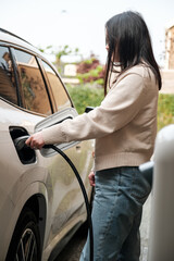 Young Central Asian woman plugging in a charging cable to charge her electric car. Sustainable and...
