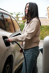 Young Central Asian woman plugging in a charging cable to charge her electric car. Sustainable and...