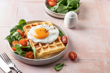 Savory breakfast with belgian waffles, fried egg and spinach, cherry tomato salad on pink tile background