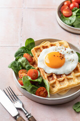 Savory waffles, fried egg and spinach, cherry tomato salad. Delicious healthy breakfast with cutlery on pink tile background
