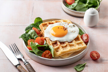 Savory waffles, fried egg and spinach, cherry tomato salad. Delicious healthy breakfast with cutlery on pink tile background