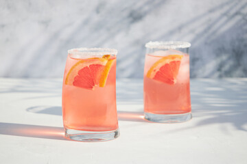 Two glasses with a fresh, cold summer grapefruit and ice cocktail, with fresh fruit slices. Red orange drink. Shadows on a sunny day