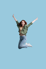 Fototapeta na wymiar Overjoyed red haired woman wearing green casual shirt, jeans, shoes jumping high looking at camera, celebration success isolated on beige background. Female having fun 