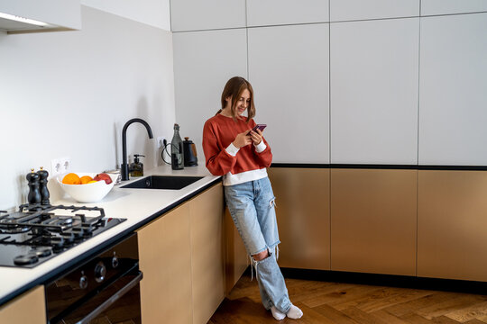 Relaxed girl stands in modern kitchen texting message on mobile phone, happy millennial teenage girl using smartphone chatting in social networks, virtual app, spends time online enjoys new mobile app