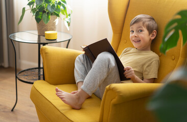A little boy sits in a chair with the book and communicates with a smart speaker