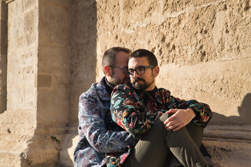 Real marriage of gay couple, sitting on a stone wall while one embraces the other from behind with very affectionate, complicit and happy attitude. Concept lgtb, lgtbiq+, couples, in love, love.