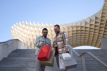 Real marriage of gay couple, standing on the stairs and with many shopping bags in their hands showing the contents to each other, happy and complicit. Concept lgtb, lgtbiq+, couples, shopping, sales