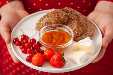 The girl in a red dress holds a plate with whole grain bread, cheese, apricot jam, red currants and strawberries. Healthy lifestyle