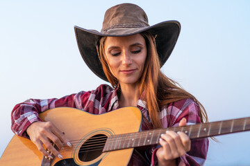 Beautiful woman playing guitar wearing a cowboy hat at dusk