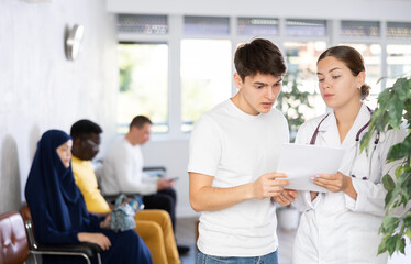 Obraz premium Polite young female doctor showing results of medical examination and explaining prescribed treatment to worried scared guy in clinic lobby