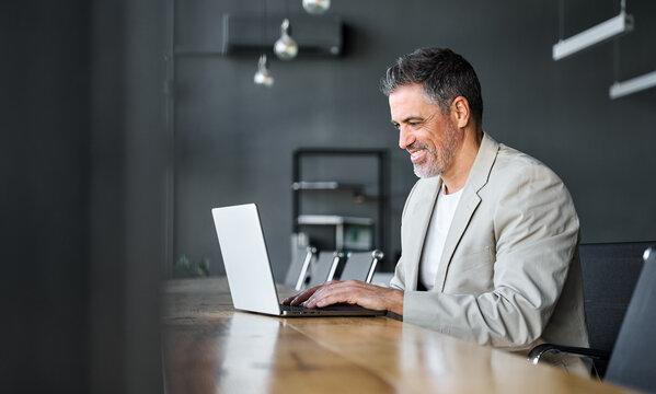 Happy busy middle aged business man ceo wearing suit sitting at desk in office using laptop. Mature businessman professional executive manager working on computer corporate technology at workplace.