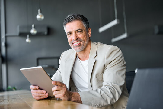 Smiling mid aged successful business man using digital tablet computer sitting in modern office. Happy mature older professional businessman entrepreneur working on technology device, portrait.