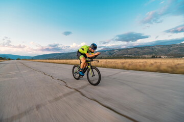  Triathlete riding his bicycle during sunset, preparing for a marathon. The warm colors of the sky provide a beautiful backdrop for his determined and focused effort.