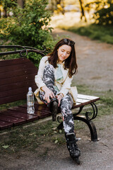 A beautiful woman, an athlete girl, sits on a wooden bench in the park in roller skates, preparing...