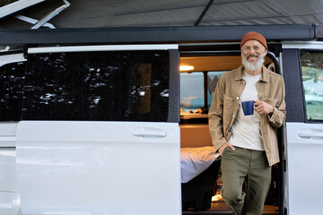 Happy older senior hipster man standing near rv camper van on vacation. Mature traveler looking at camera, holding drinking coffee waking up in the morning in camping tourism nature park.