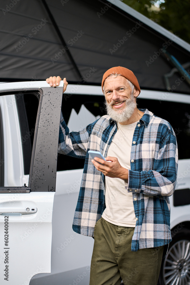 Wall mural Happy elder man standing near rv camper van on vacation using mobile phone. Old mature active traveler holding smartphone in camping nature park advertising campervan booking rental service. Vertical