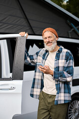 Happy elder man standing near rv camper van on vacation using mobile phone. Old mature active...