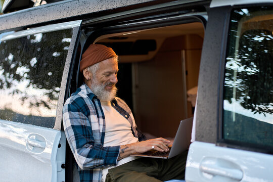 Happy older man sitting in rv camper van using laptop. Smiling mature active traveler holding computer on lap remote working online and enjoying vanlife, freedom, resting in outdoor camping.