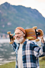 Active cool happy bearded old hipster man standing in nature park holding skateboard. Mature...