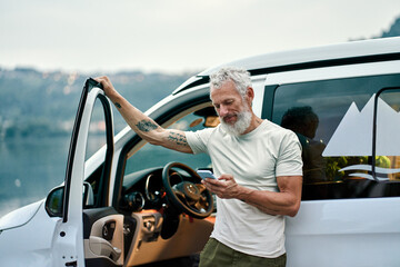 Mature tattooed man standing near rv camper van on vacation using mobile phone. Smiling mature...