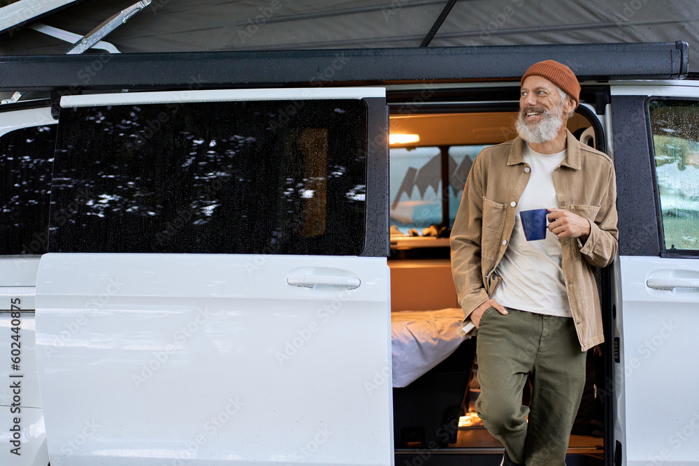 Wall mural Active older happy hipster man standing near rv camper van on vacation. Mature traveler looking away enjoying view, holding drinking coffee waking up in the morning in camping tourism nature park.