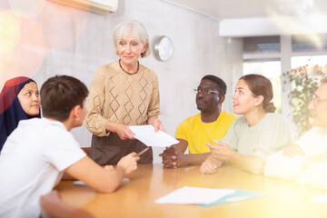 Obraz premium Group of interested people on various ages and nationalities communicating in foreign language speaking club with positive elderly female tutor..