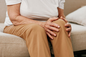 Elderly woman severe pain in her leg sitting on the couch, health problems in old age, poor quality of life. Grandmother with gray hair holds on to her sore knee, problems with joints and ligaments. - obrazy, fototapety, plakaty