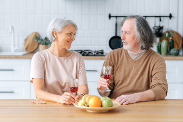 Obraz na płótnie Canvas Middle aged caucasian spouses, sit at home in the kitchen, hold glasses of red wine in their hands, celebrate an important date, anniversary, look at glasses, smile joyfully. Happy pensioners