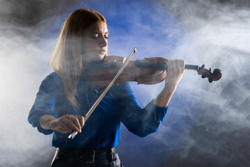 Beautiful blond girl playing violin. Female violinist against dark blue background with smoke.