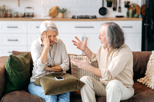 Mature caucasian couple, pensioners, sit on the sofa in the living room, quarrel with each other, shout, swear, experience irritation and indignation, disagree with each other, gesticulate with hands