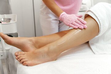 Fototapeta na wymiar Close up photo of applying cream. Cosmetological procedures in a beauty salon. A woman beautician in a pink gloves applies a moisturizer to her female patient's leg after epilation, leg hair removal