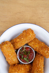 Top view of fried mozzarella cheese fingers with a red spicy dipping sauce, in a white bowl on the wooden table.