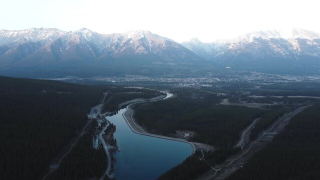 Orbiting aerial drone shot of a lake in front of snowy and beautiful Canadian peaks with Canmore, Alberta nestled between the two