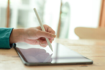 Close up image of a woman is using stylus pen and empty screen tablet at the wooden table.