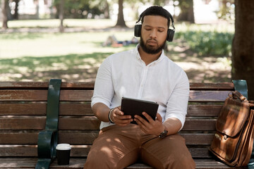 Fototapeta premium Business man, headphones and outdoor with a tablet on a park bench for a break with internet. Male digital nomad and person in nature while streaming and listening to music on a audio app to relax