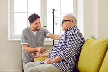Young friendly smiling nurse man helping a senior grey-haired patient sitting on the couch at home receiving iv drip infusion and injection vitamin therapy in his blood. Nursing help concept. - obrazy, fototapety, plakaty