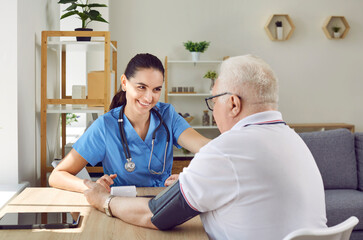 Obraz premium Friendly young nurse measures blood pressure of older male patient in nursing home. Female caregiver sits at table and uses electric tonometer while examining older gray-haired man. Concept of nursing