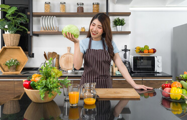 Young asian housewife dressed in an apron, hold fresh lettuce with one hand. The kitchen counter full of various kinds of vegetables.