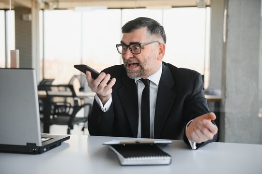 stressed handsome businessman working at desk in modern office shouting at laptop screen and being angry about financial situation, jealous of rival capabilities, unable to meet client needs