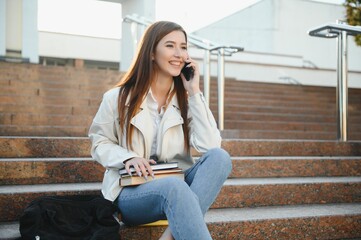 Portrait of a cheerful young girl student with backpack sitting on steps outdoors, reading book