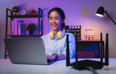 Happy young Asian woman using laptop social media messages on the table for modem to high speed internet via LAN cable in modern home office at night.