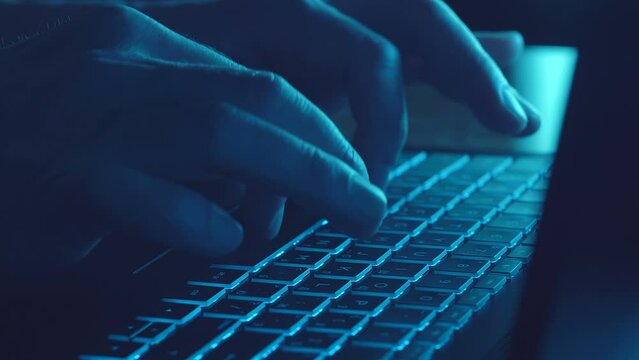 Man's Hands Typing On A Black Laptop With Illuminated Keyboard Lying On A Desktop On Dark. Closeup. Shallow Depth Of Field