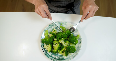 Woman eating broccoli on the kitchen with knife and fork.