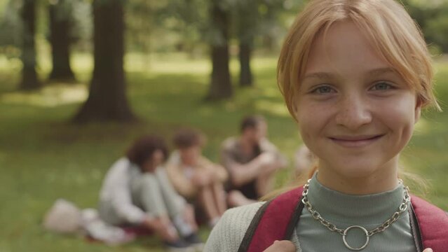 Medium closeup portrait of smiling Caucasian girl with cute dimples on her cheeks looking at camera while standing in summer park during outdoor lesson with classmates and teacher, recreation, leisure