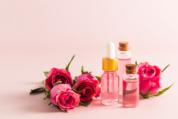 Serum with rose petals and rose water in glass bottles against a background of live pink roses. A fashionable cosmetic product for young skin.