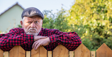 Ein Mann mit Mütze steht grimmig hinter einem Zaun und blickt zornig zu den Nachbarn - obrazy, fototapety, plakaty