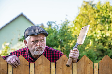 Ein aggressiver Mann hält drohend eine Waffe in der Hand. Er blickt grimmig und steht hinter einem Zaun. Hinter ihm sind Büsche und ein Gebäude zu sehen. - obrazy, fototapety, plakaty