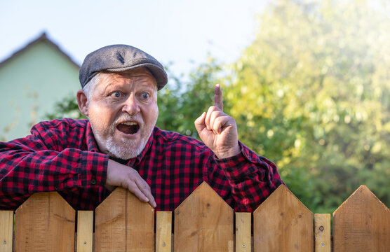 Ein Mann mit Mütze steht grimmig hinter einem Zaun und blickt zornig zu den Nachbarn
